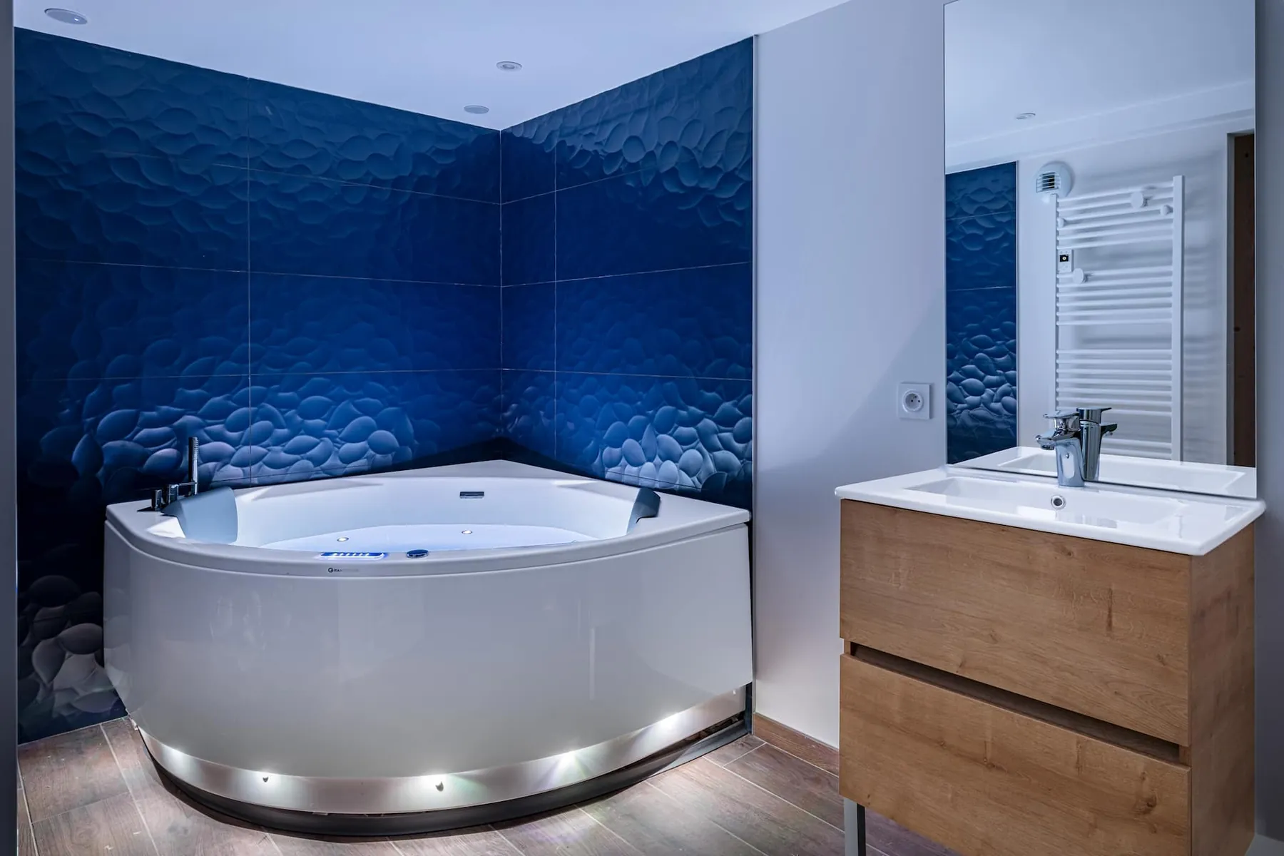 La baignoire balnéo d'angle devant son mur bleu nuit texturé, au rez-de-chaussée de la maison